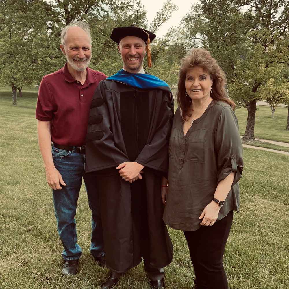 A middle aged man in graduation attire stands between his parents.
