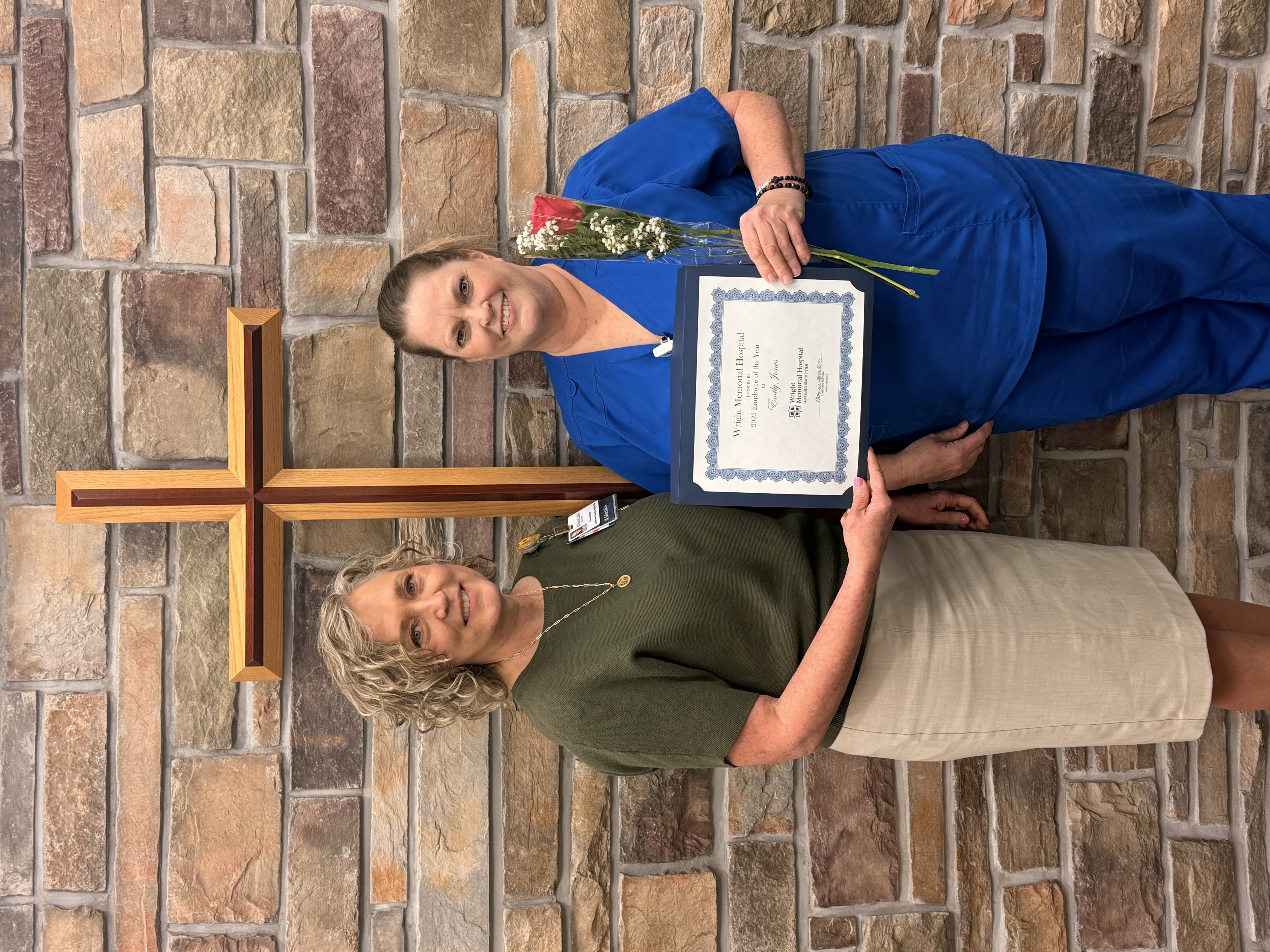 Emily Jones wears blue scrubs (right), accepting award from hospital administrator Catherine Hamilton, both standing across a brick wall 