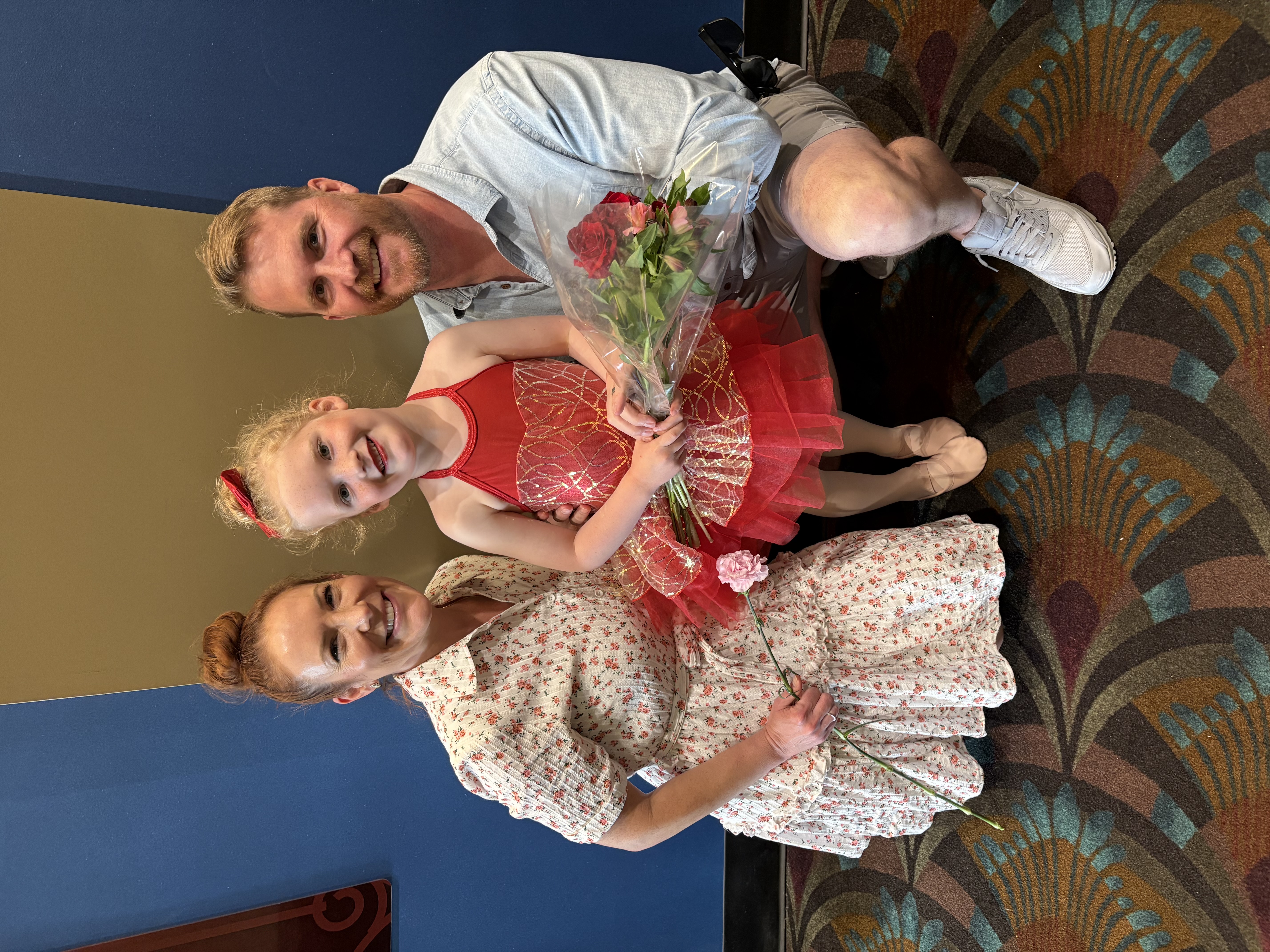 Jim (left) and his wife sit next to their daughter (middle) who is wearing a red dress & carrying roses.