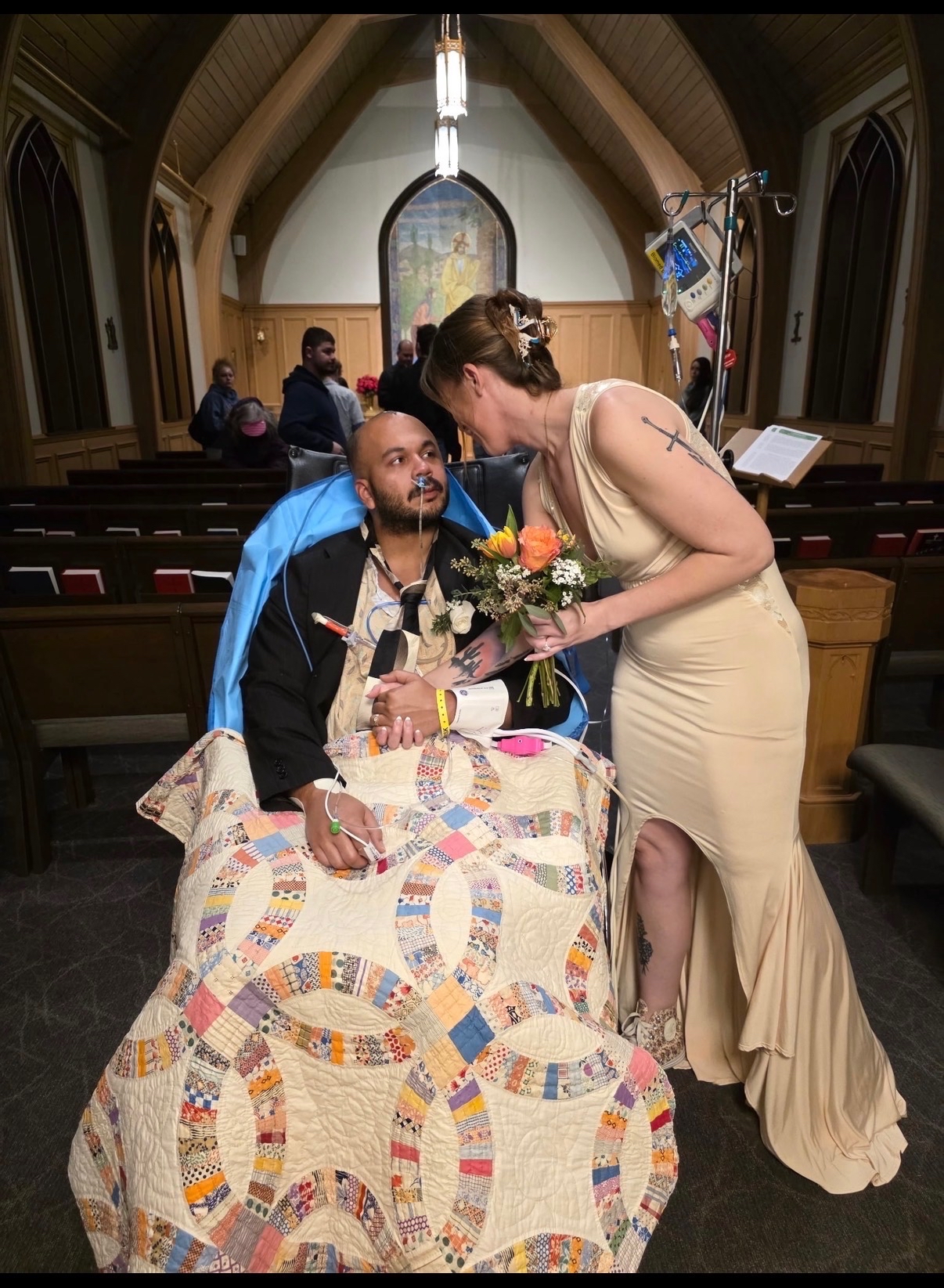 Justin (left) seated on a wheelchair with blanket on lower body and nasal tube, with his new wife Nicole bent near him, holding a peach flower bouquet and wearing a beige sleeveless dress at the chapel